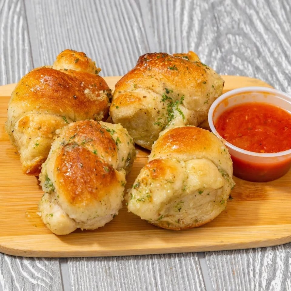 Golden-brown garlic knots with herbs and parmesan, served with a side of marinara sauce on a wooden board.