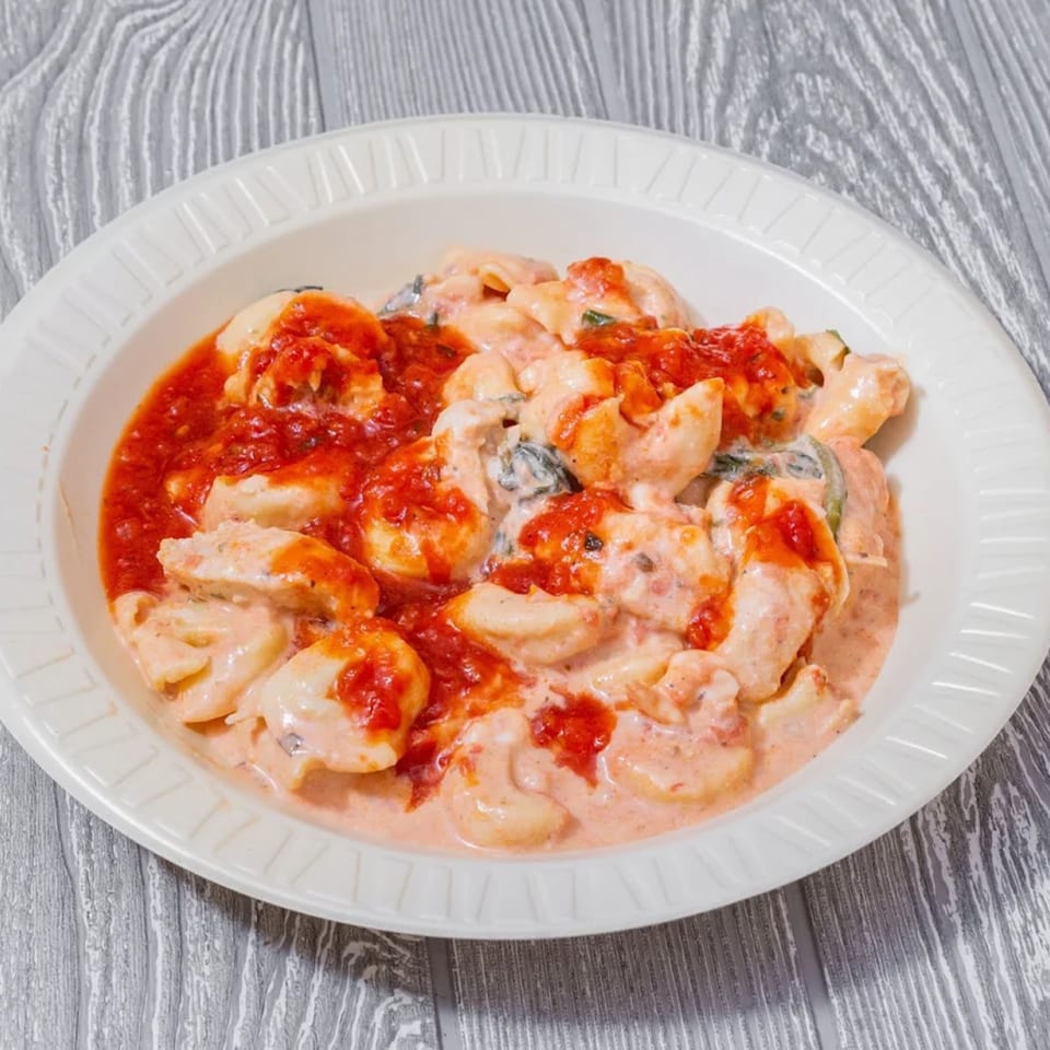 A white bowl filled with tortellini pasta in a creamy pink sauce, topped with red marinara.
